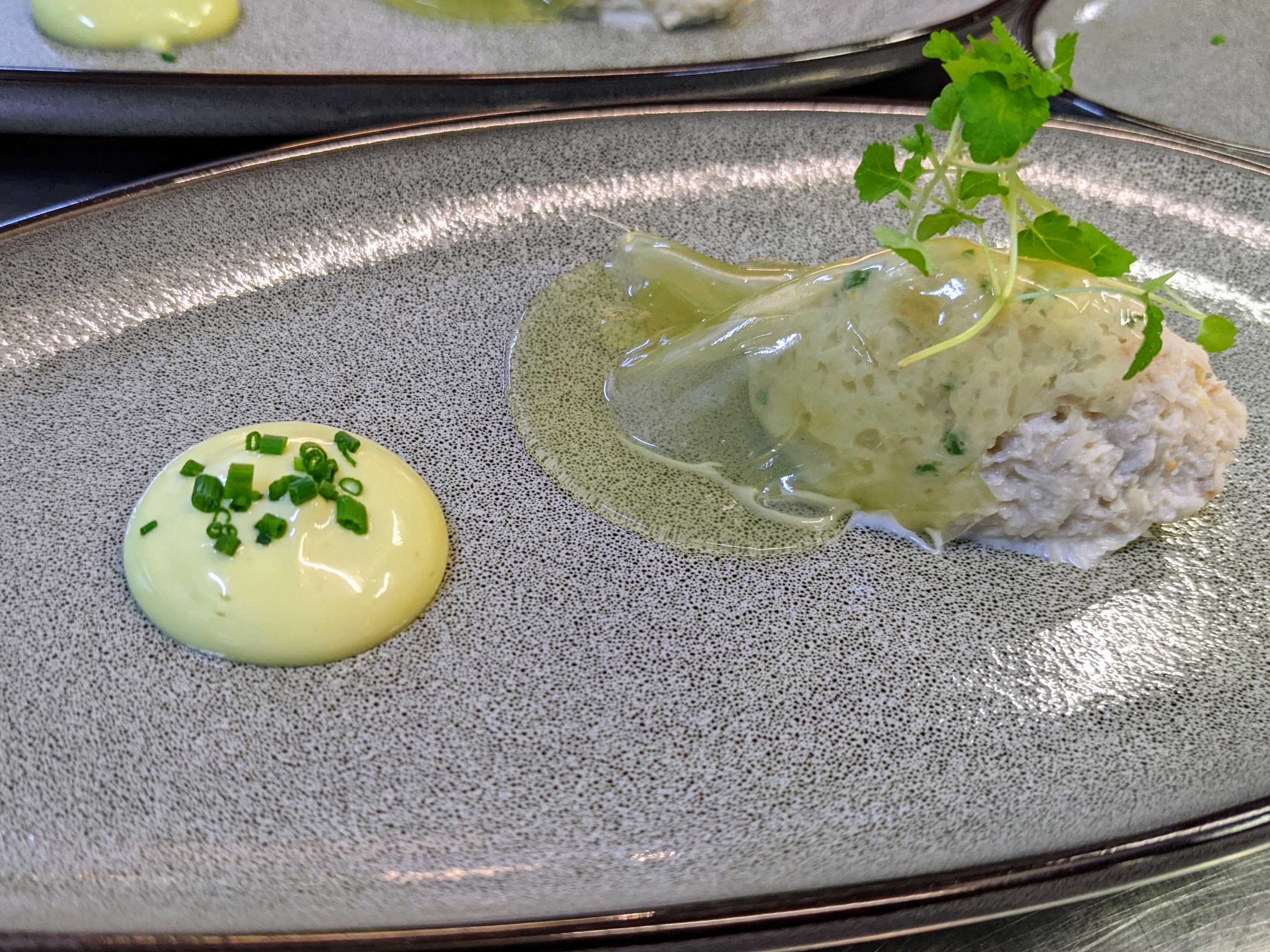 A grey plate containing a green emulsion topped with chives, and a spoon full of dressed white crab with cucumber jelly draped over it, topped with micro lemon balm.
