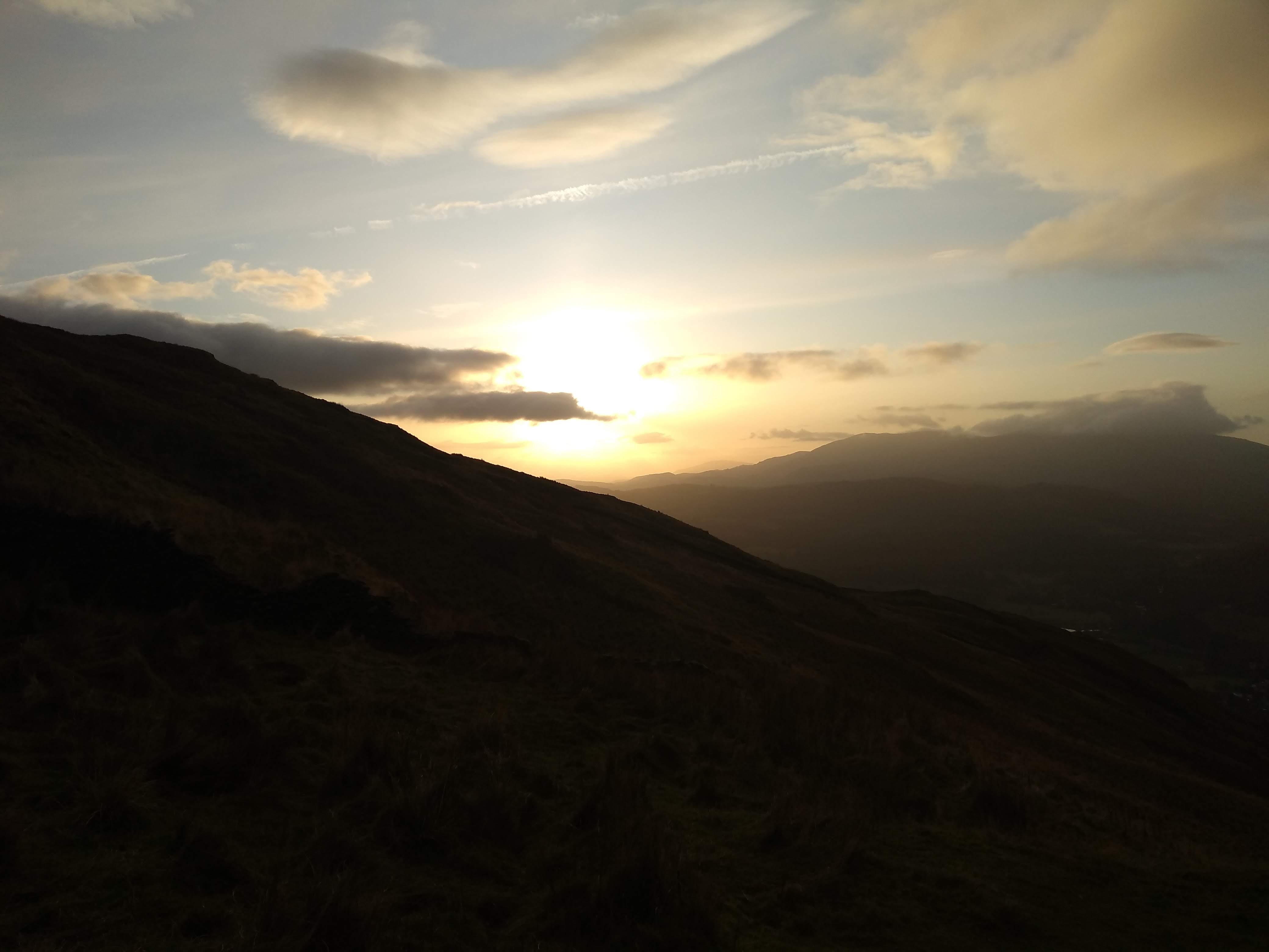 A landscape photo of the sun through the clouds casting rays over rolling hills.